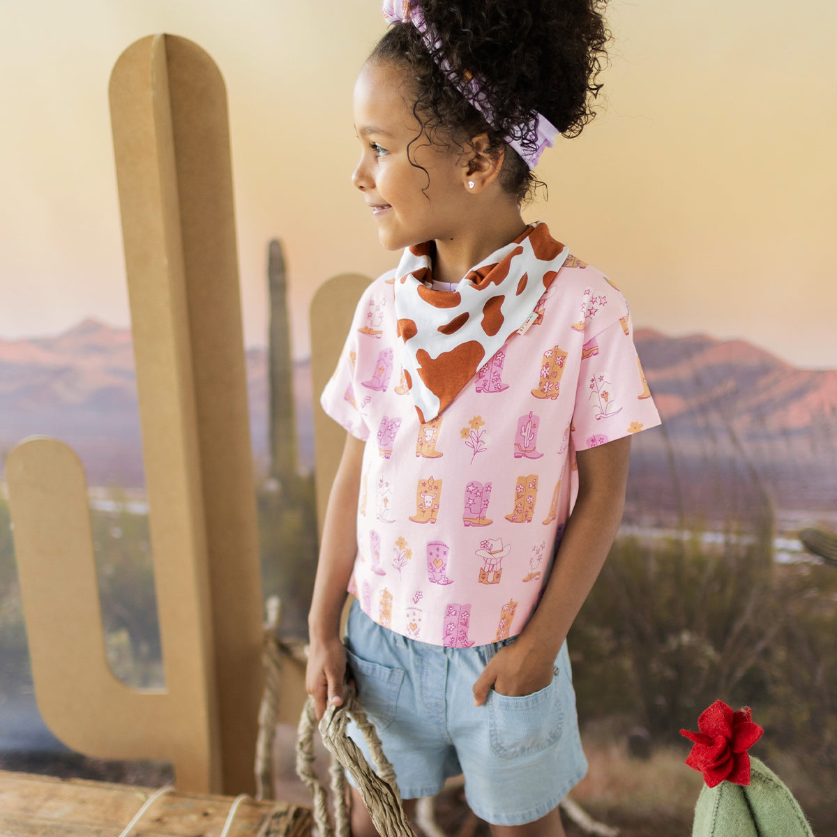 Child wearing a pink shirt with cowboy print and denim shorts in front of a desert landscape backdrop.