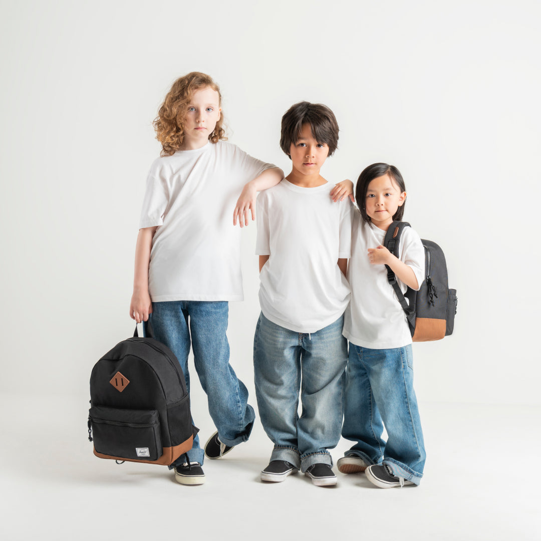 Three children standing together in a studio wearing plain white T-shirts and blue jeans, with two of them carrying black backpacks. The background is a clean white studio backdrop.