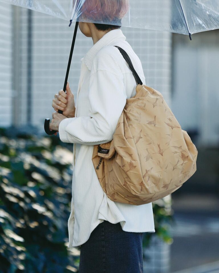 Person holding a beige Shupatto bag and transparent umbrella outdoors