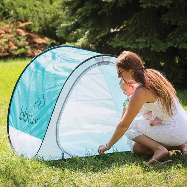Woman setting up a blue and white baby tent in a grassy area with trees in the background.