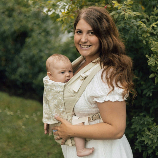Woman holding a baby in a sling outdoors with greenery in the background